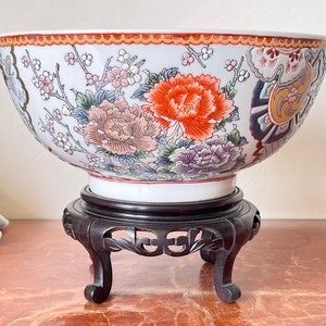 May include: A large white porcelain bowl with a floral design in shades of red, orange, purple, and blue. The bowl is sitting on a dark wooden stand with three legs.