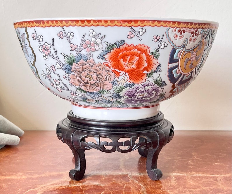 May include: A large white porcelain bowl with a floral design in shades of red, orange, purple, and blue. The bowl is sitting on a dark wooden stand with three legs.
