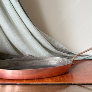 May include: A copper-colored frying pan with a long, angled handle. The pan has a wide, shallow bowl and is set on a brown surface. A light green fabric is draped in the background.