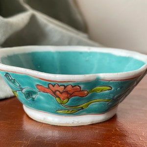 Puede incluir: Un peque&ntilde;o cuenco de cer&aacute;mica con bordes festoneados, con un esmalte turquesa y un dise&ntilde;o floral. El cuenco presenta flores y hojas rojas, naranjas y verdes.