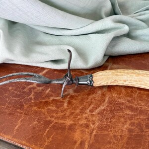May include: An antique meat fork with a light-colored antler handle and a silver-toned metal head. The fork has two tines and a curved hook. The handle is attached to the head with a decorative silver-toned metal collar. The fork rests on a brown leather surface.