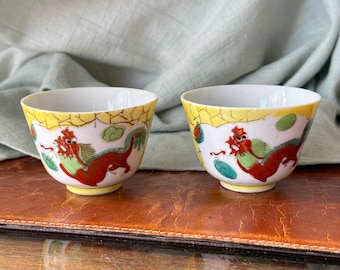 May include: Two small, white porcelain bowls with a yellow rim and a dragon design. The dragons are red with green accents. The bowls are on a brown surface.