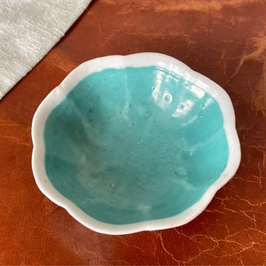 Puede incluir: Un peque&ntilde;o cuenco de cer&aacute;mica blanca con un borde festoneado y un interior azul turquesa. El cuenco est&aacute; sobre una superficie de cuero marr&oacute;n.