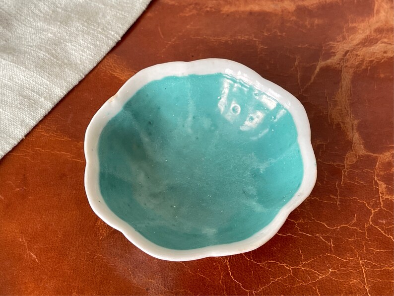 Puede incluir: Un peque&ntilde;o cuenco de cer&aacute;mica blanca con un borde festoneado y un interior azul turquesa. El cuenco est&aacute; sobre una superficie de cuero marr&oacute;n.