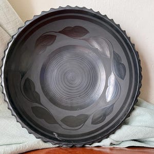 May include: A dark gray ceramic bowl with a spiral design in the center and leaf patterns around the rim. The bowl has a textured, scalloped edge and is displayed on a wooden surface.