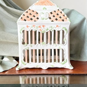 May include: A white ceramic birdcage-shaped decorative piece with a lattice roof and vertical bars. The cage features peach and green accents, including floral designs and a decorative handle. The piece is displayed on a reflective surface.