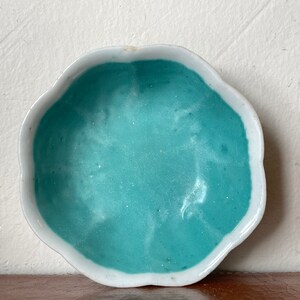 Puede incluir: Un peque&ntilde;o plato de cer&aacute;mica blanca con un borde festoneado. El interior del plato est&aacute; pintado de un azul turquesa claro.