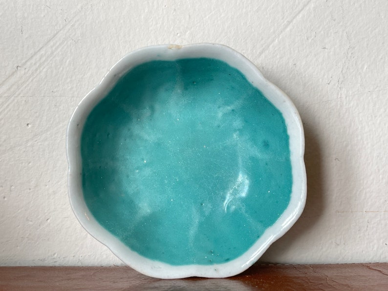 Puede incluir: Un peque&ntilde;o plato de cer&aacute;mica blanca con un borde festoneado. El interior del plato est&aacute; pintado de un azul turquesa claro.