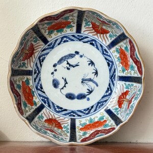 May include: A decorative ceramic plate with a scalloped edge. The plate features a white center with a blue bird and floral design. The rim has alternating red and blue floral patterns. The plate's edge is a light tan color.