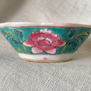 Puede incluir: Un peque&ntilde;o cuenco de cer&aacute;mica blanca con un borde festoneado. El cuenco est&aacute; decorado con un fondo azul verdoso y una flor rosa con hojas verdes.