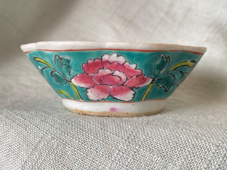 Puede incluir: Un peque&ntilde;o cuenco de cer&aacute;mica blanca con un borde festoneado. El cuenco est&aacute; decorado con un fondo azul verdoso y una flor rosa con hojas verdes.