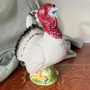 May include: A ceramic turkey figurine with a white body, black and white tail feathers, and a red head. The turkey has a blue beak and yellow feet, standing on a green base. The figurine is likely a decorative item.
