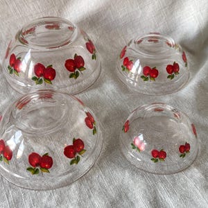 May include: Four clear glass bowls of varying sizes, each adorned with a repeating pattern of red apples with green leaves. The bowls are arranged on a light-colored, textured surface, showcasing a vintage kitchenware set.