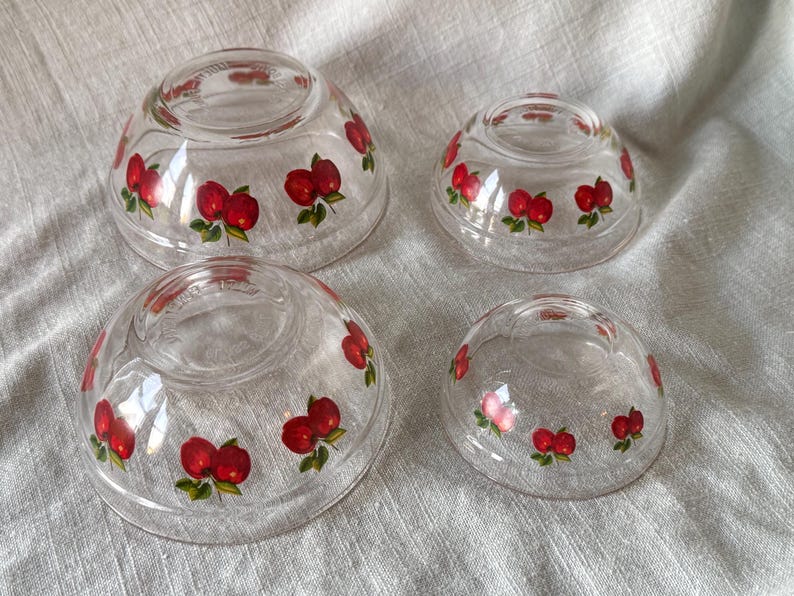 May include: Four clear glass bowls of varying sizes, each adorned with a repeating pattern of red apples with green leaves. The bowls are arranged on a light-colored, textured surface, showcasing a vintage kitchenware set.