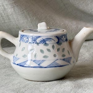 May include: Small white ceramic teapot with a lid, handle, and spout. The teapot is decorated with blue and light green leaf and geometric patterns. The lid has a small square handle.