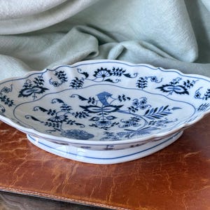 May include: Oval, white ceramic serving dish with a blue floral pattern. The dish has a scalloped edge and a raised base. The blue design features stylized flowers and foliage. The dish is on a brown surface.