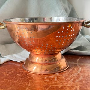 May include: A polished copper colander featuring a wide, circular base and two curved brass handles on opposite sides. The bowl is perforated with numerous small, evenly spaced holes and has a smooth, reflective metallic finish throughout.