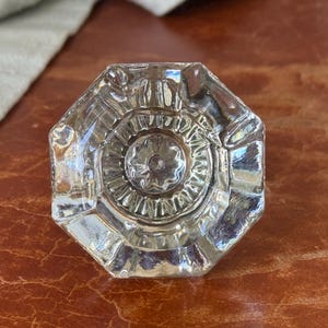 May include: A clear, octagonal glass cabinet knob featuring a faceted design with a central starburst pattern. The knob has a multi-sided geometric shape with reflective surfaces and a circular center detail, providing a transparent appearance.