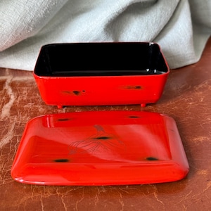 May include: A red lacquered box with a black interior and a matching lid. The lid features a black illustration. The box is rectangular and sits on small feet. The box and lid are on a brown surface.