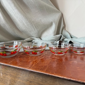 May include: Set of four clear glass bowls with a red cherry design around the rim. The bowls are graduated in size, perfect for serving snacks or desserts. The bowls are arranged on a brown surface.