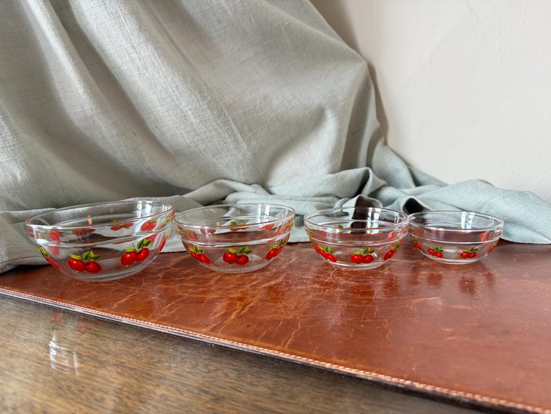May include: Set of four clear glass bowls with a red cherry design around the rim. The bowls are graduated in size, perfect for serving snacks or desserts. The bowls are arranged on a brown surface.