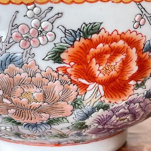 May include: A white ceramic bowl with a floral design. The bowl features pink, orange, and blue flowers with green leaves. The bowl has a gold rim.
