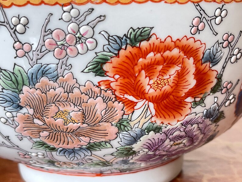 May include: A white ceramic bowl with a floral design. The bowl features pink, orange, and blue flowers with green leaves. The bowl has a gold rim.
