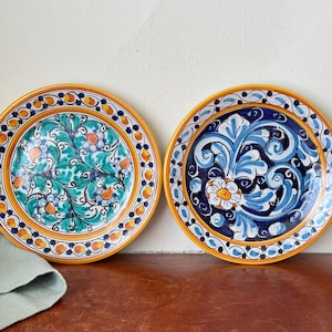 May include: Two decorative ceramic plates with hand-painted floral designs. One plate features a teal and orange floral pattern, while the other showcases a blue and white design. Both plates have a yellow border with decorative accents.