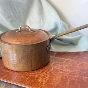 May include: A round copper saucepan with a matching lid and a long, slender brass handle. The exterior of the pot and the lid feature a hammered texture, showing a warm, metallic orange-brown finish with visible signs of natural patina.