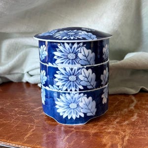 May include: A three-tiered, blue and white ceramic container with a lid. The container and lid are decorated with a floral pattern of white daisies and leaves. The container is likely used for food storage or display.