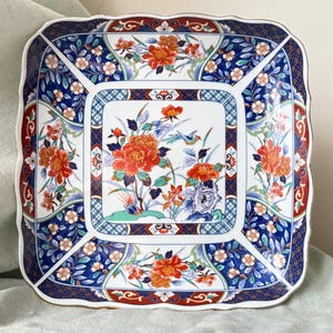 May include: Square ceramic decorative plate with a scalloped edge. The plate features a floral design with orange, blue, and white flowers, and a gold trim. The plate has a white center with a floral and bird design.