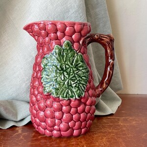May include: A pink ceramic pitcher with a grape design. The pitcher has a brown handle shaped like a grapevine.