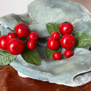 May include: Two sprigs of faux holly with red berries and green leaves on a light blue velvet background.