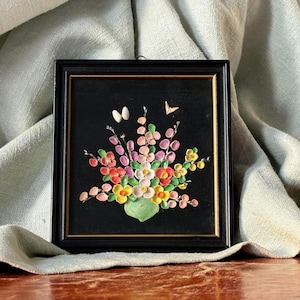 May include: A framed floral artwork with a black background. The artwork features a bouquet of colorful flowers in shades of pink, red, yellow, and white. Two white ovals and a small butterfly are above the flowers. The frame is black.