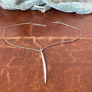 May include: A silver necklace with a long, thin pendant that resembles a blade. The necklace is on a brown leather surface.