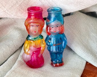 May include: Two vintage glass figurines, one in pink, yellow, and red, the other in blue and red. The figures are shaped like people with detailed faces and clothing. They are likely Christmas ornaments or decorative items.