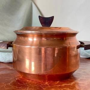 May include: Copper pot with wooden handles and a wooden lid knob. The pot is shiny and has a rounded shape.