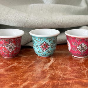 May include: Three small, decorative porcelain cups with a floral design. The cups have a white interior and a colorful exterior. One cup is red, one is turquoise, and one is red with a white floral design. The cups are likely for tea or espresso.