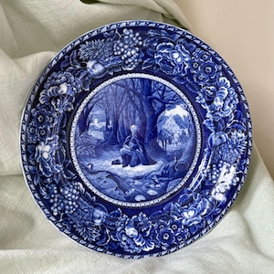May include: A round ceramic plate featuring a deep blue and white transferware pattern. The center depicts a figure kneeling in a wooded landscape, while the wide border is decorated with detailed illustrations of various fruits and flowers.