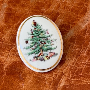 May include: Oval-shaped Christmas ornament with a gold rim. The ornament features a detailed illustration of a decorated Christmas tree with presents underneath. The tree is green with red, white, and gold ornaments. A festive holiday decoration.