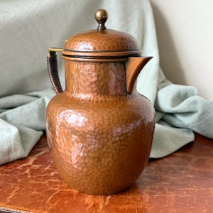 May include: A hammered copper coffee pot with a lid and a spout. The pot has a dark brown handle and a brass-colored knob on top. The copper has a warm, reddish-brown hue and a textured surface. The pot is likely vintage or antique.