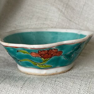 Puede incluir: Un peque&ntilde;o cuenco de cer&aacute;mica turquesa con bordes festoneados y un dise&ntilde;o floral. El cuenco tiene un borde blanco y una base blanca. El dise&ntilde;o floral presenta flores rojas y verdes.
