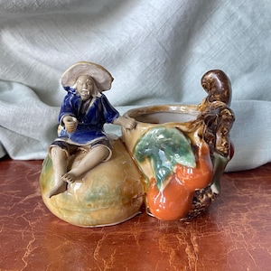 May include: Ceramic planter shaped like two gourds with a small figurine of a man sitting on top. The planter is decorated with green, brown, and orange paint.