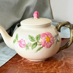 May include: A cream-colored ceramic teapot featuring a hand-painted floral design of pink roses with green leaves. The teapot has a matching lid topped with a small pink rosebud finial and a dark green, textured handle shaped like a twisted branch.