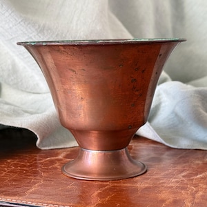 May include: A vintage copper cup with a flared rim and a pedestal base. The cup has a rich, reddish-brown patina with some darker spots, suggesting age and use. The rim has a greenish hue. The cup sits on a brown leather surface.