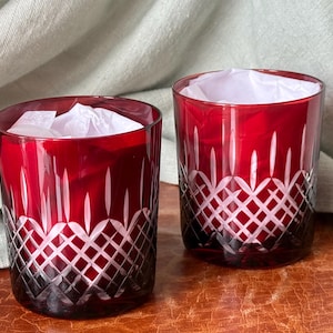 May include: Two ruby red drinking glasses with a cut crystal design. The glasses are cylindrical with a geometric pattern near the base and vertical lines extending upwards. Each glass contains white tissue paper.