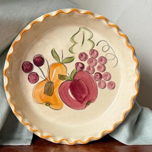 May include: A cream-colored pie plate with a scalloped orange rim. The plate is decorated with hand-painted fruit: a red apple, an orange pear, purple grapes, and cherries. The plate is on a wooden surface with a blue cloth.