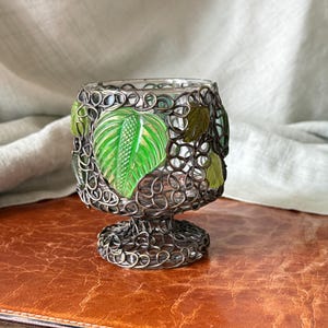 May include: A clear glass votive candle holder with a decorative metal filigree overlay. The holder features green and olive-colored leaf accents. The base and rim have a detailed, circular pattern. The candle holder sits on a brown surface.