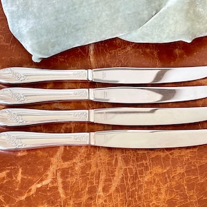 May include: Four silver dinner knives with ornate floral designs on the handles. The knives are arranged in a row on a brown leather surface.