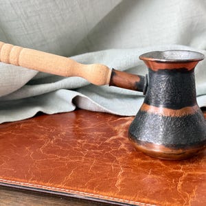 May include: A small, hammered copper coffee pot with a wooden handle. The pot has a black body with a copper band and rim. The handle is light-colored wood. The pot is designed for making Turkish or Greek coffee.